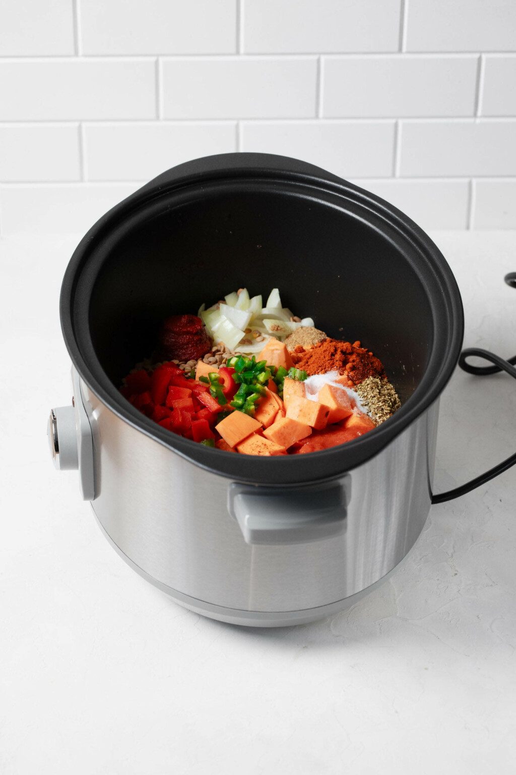 A silver slow cooker is filled with vegetables and lentils for making a batch of chili.