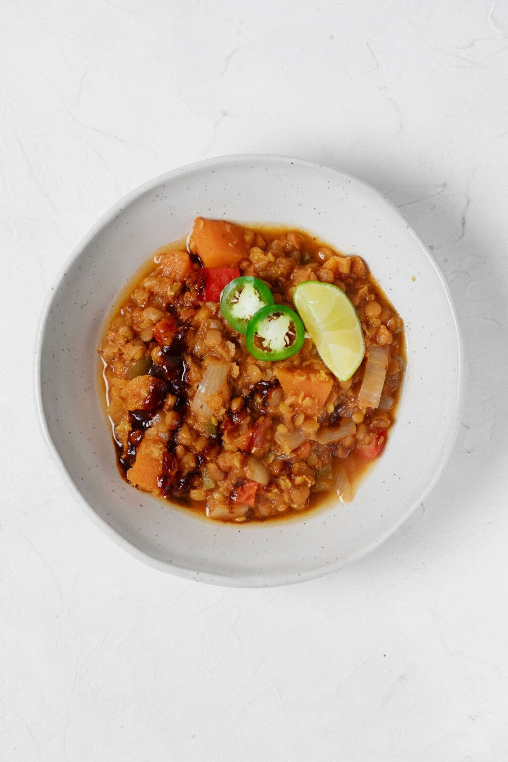 An angled image of a shallow white bowl, which is being used to hold a plant-based lentil chili and garnishes.