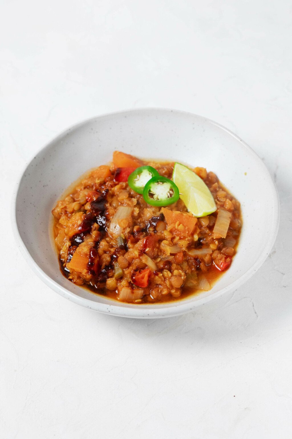 An angled image of a shallow white bowl, which is being used to hold a plant-based lentil chili and garnishes.