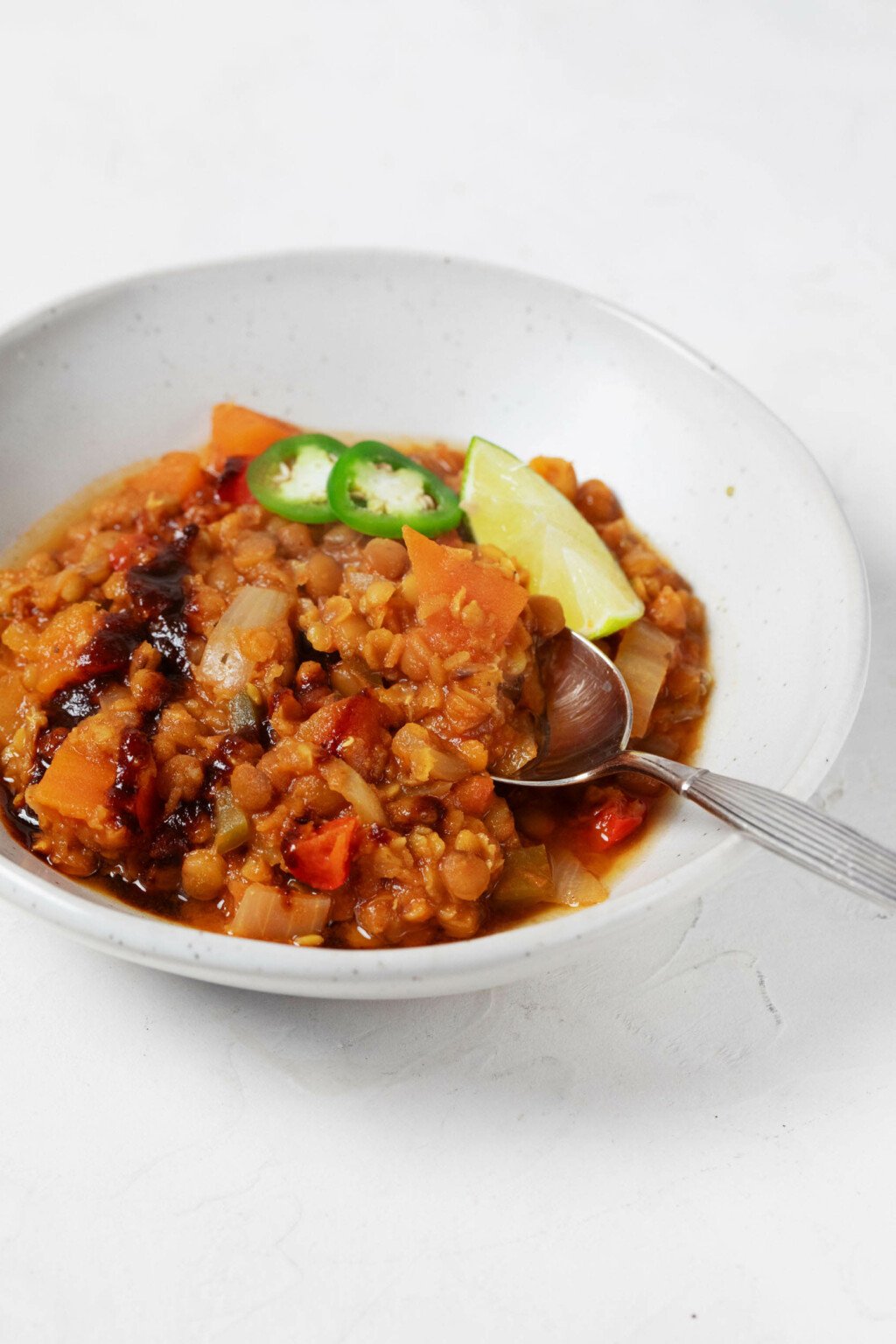 An angled image of a shallow white bowl, which is being used to hold a plant-based lentil chili and garnishes.