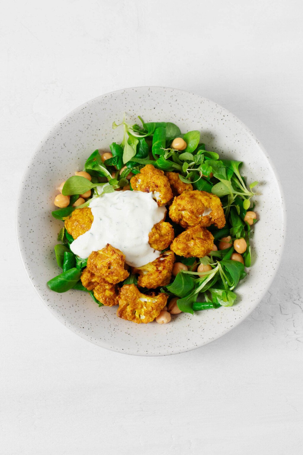 An overhead image of a bed of greens topped with tandoori spiced cauliflower, chickpeas, and a creamy sauce.