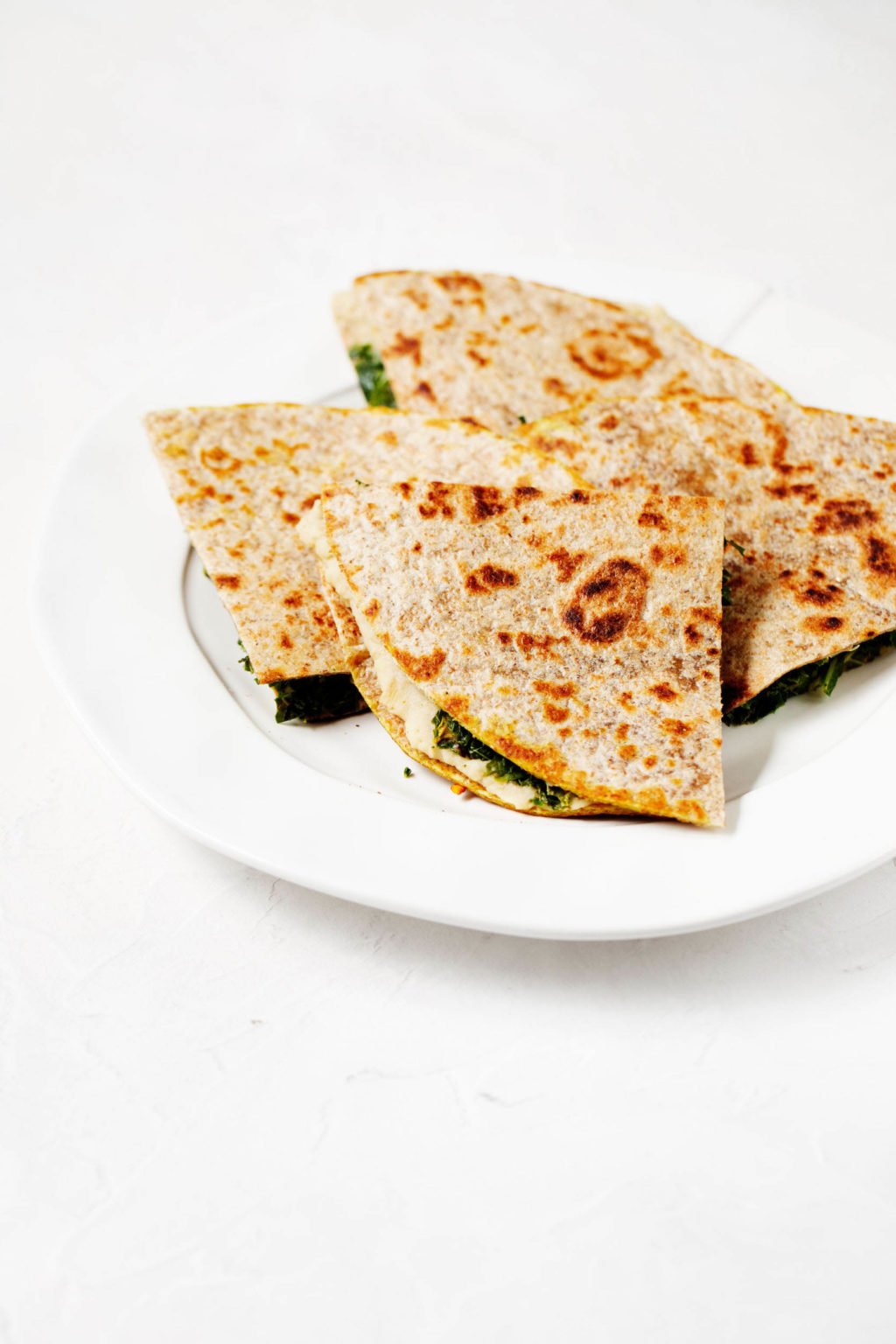 A round, rimmed white plate has been stacked with triangular wedges of white bean artichoke wraps, which also contain bright green kale.