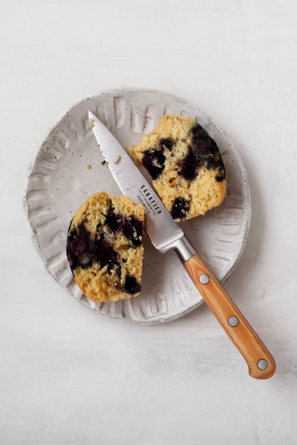 An overhead image of a fruit muffin that has just been sliced in half. The cut halves are on a small dessert plate with a paring knife.