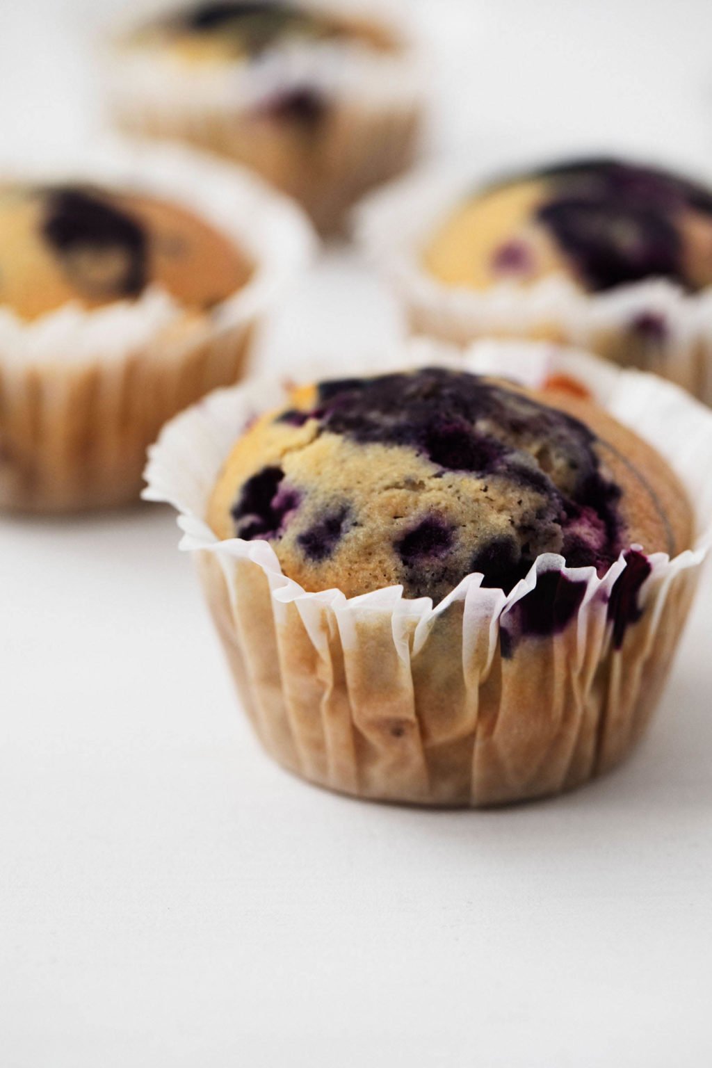 A few corn muffins, filled with juicy blueberries, are resting in muffin liners on a white surface.