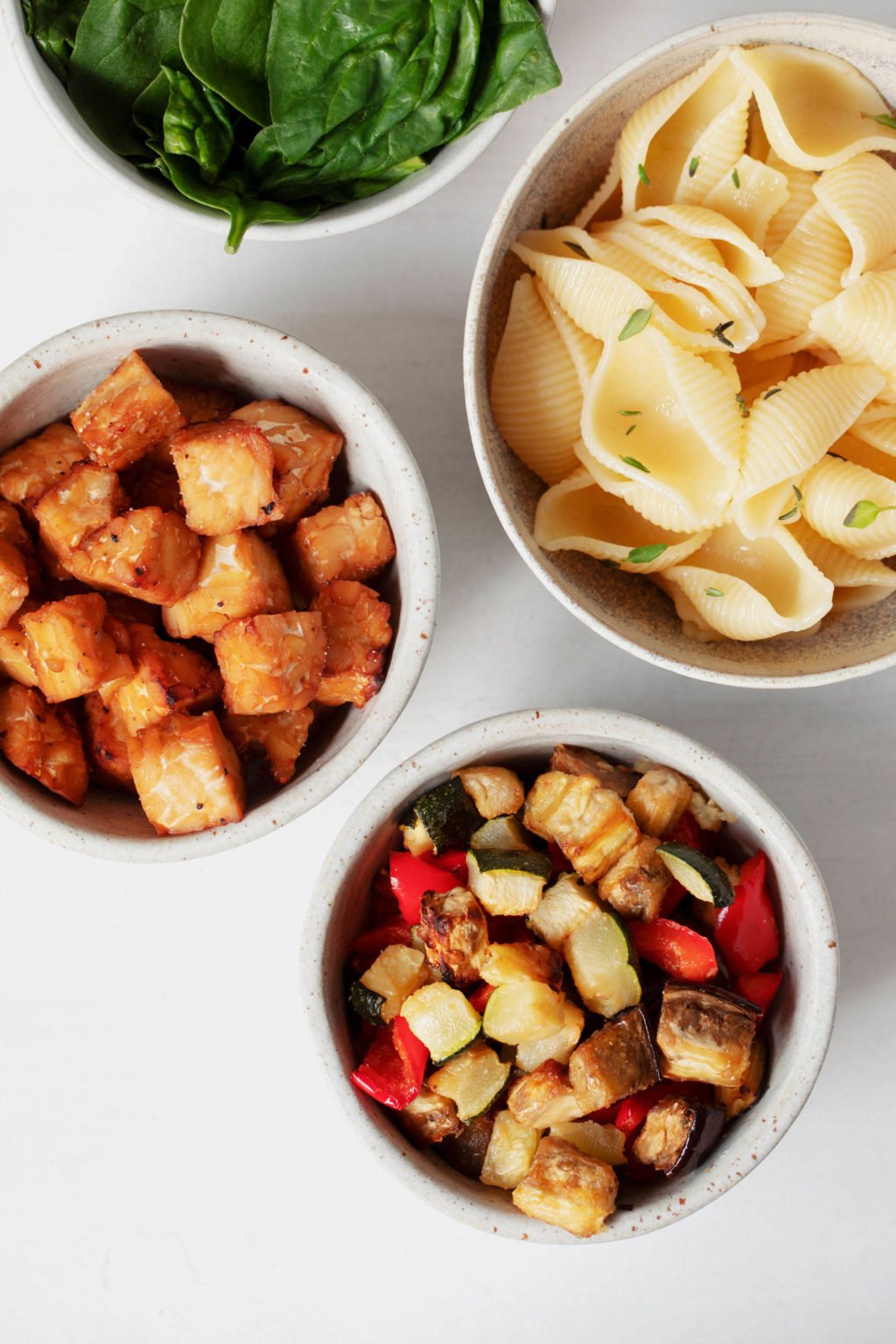 A few small, white bowls are filled with the ingredients needed to make a vegetable pasta salad.