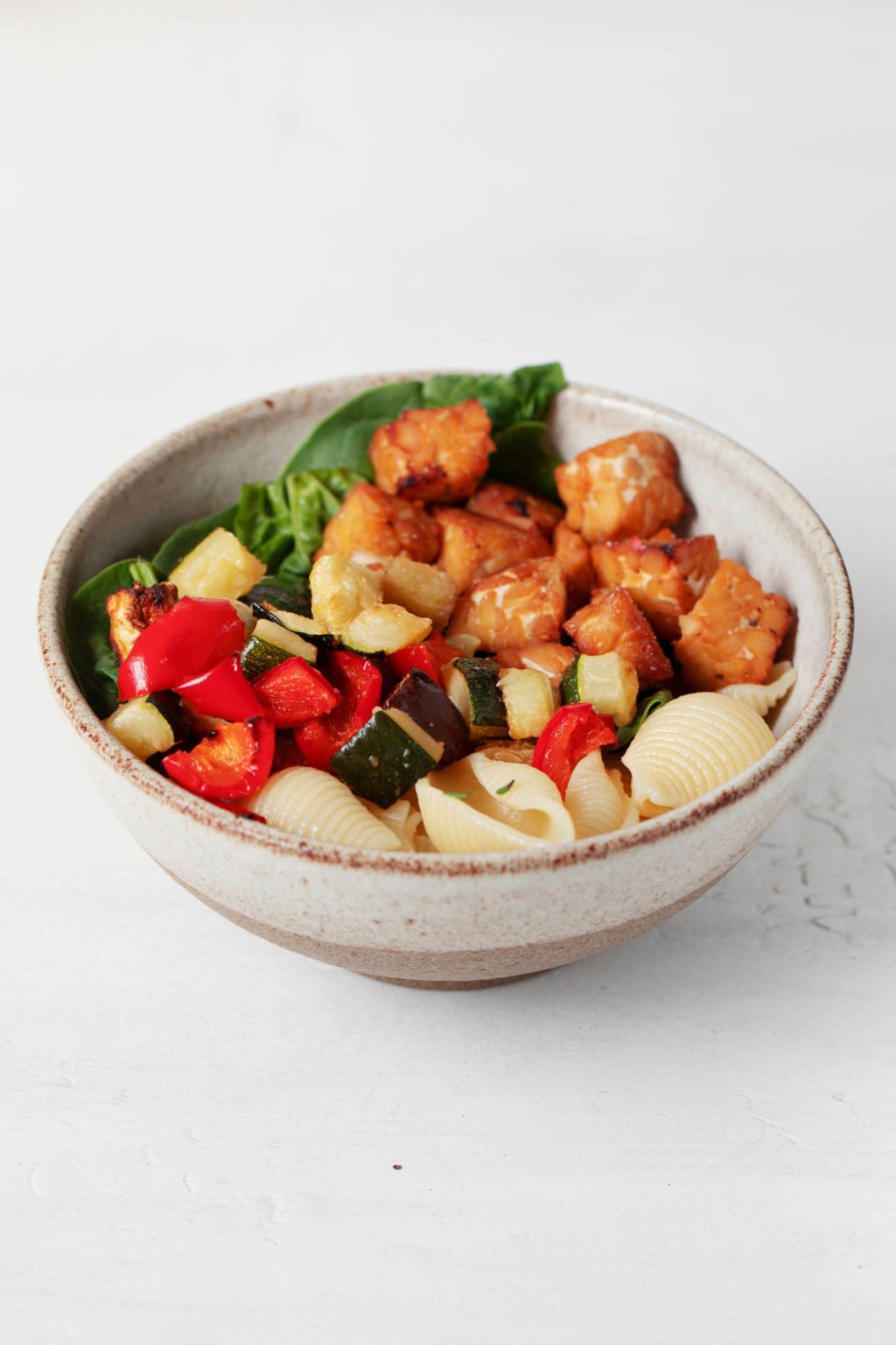 A small, white and beige bowl is resting on a white surface. The bowl is filled with cooked pasta and colorful mix-ins.