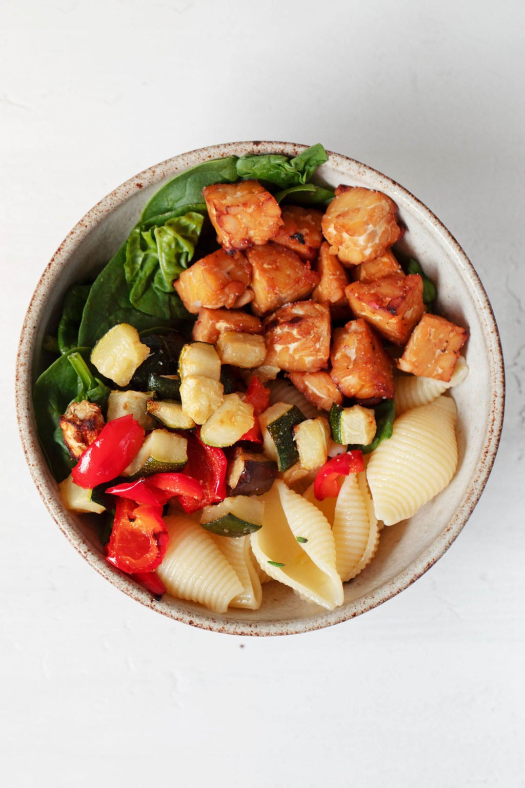 An overhead image of a serving dish that holds roasted tempeh cubes, spinach, and roasted vegetables. The colorful mixture is set against a white backdrop.