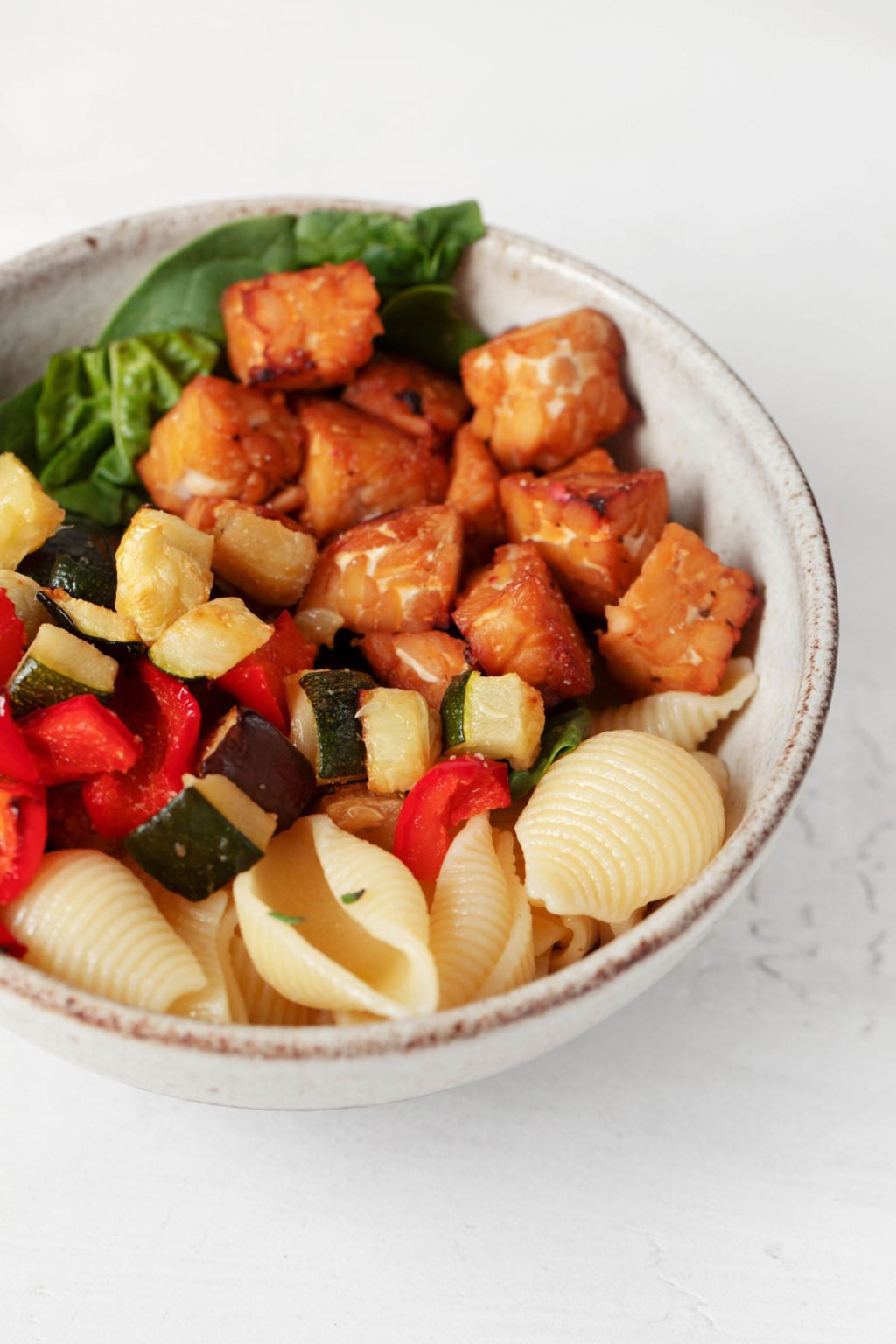 A round, white ceramic bowl has been filled with a colorful tempeh vegetable pasta dish. It rests on a white surface.