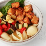 A round, white ceramic bowl has been filled with a colorful tempeh vegetable pasta dish. It rests on a white surface.