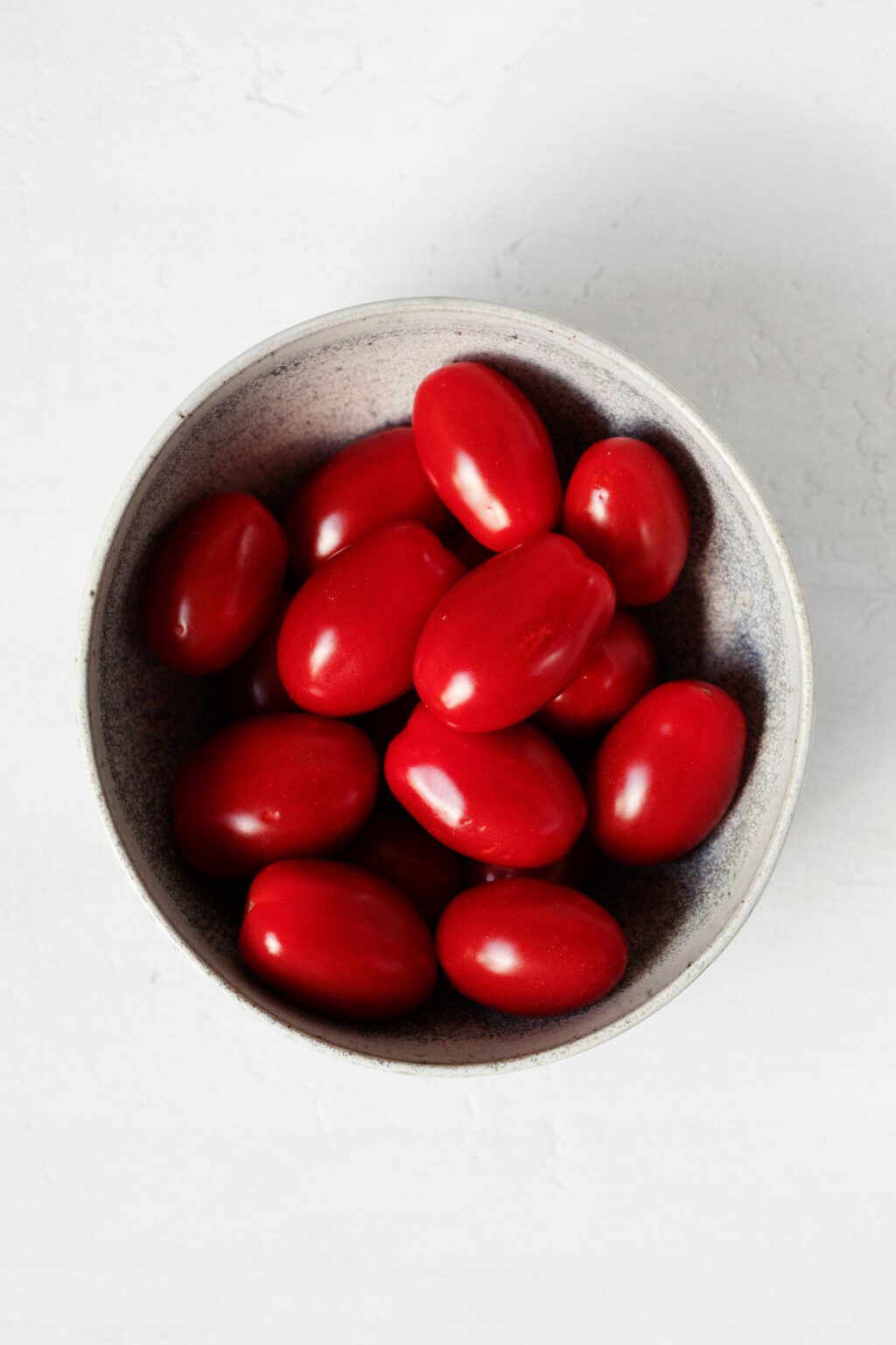 A small white bowl of bright red grape tomatoes rests on a white surface.