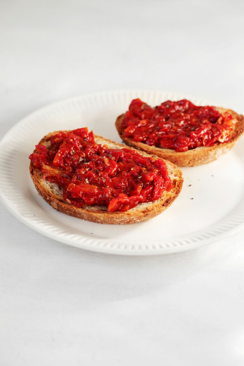 Two oval-shaped slices of toast are topped with a bright red cherry tomato jam.