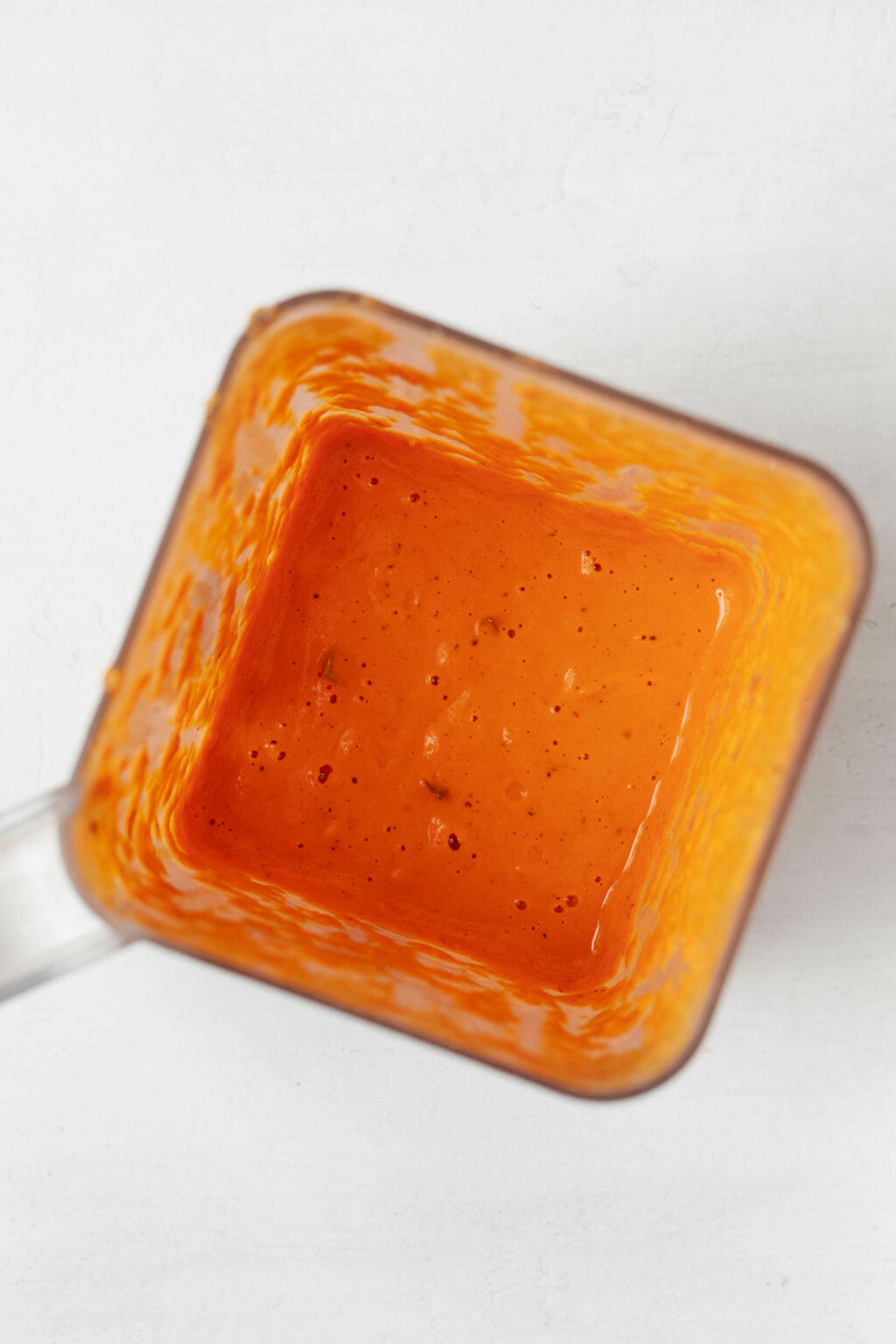 An overhead image of a blender container, which has a square-shaped mouth. The container holds an orange-colored sauce.