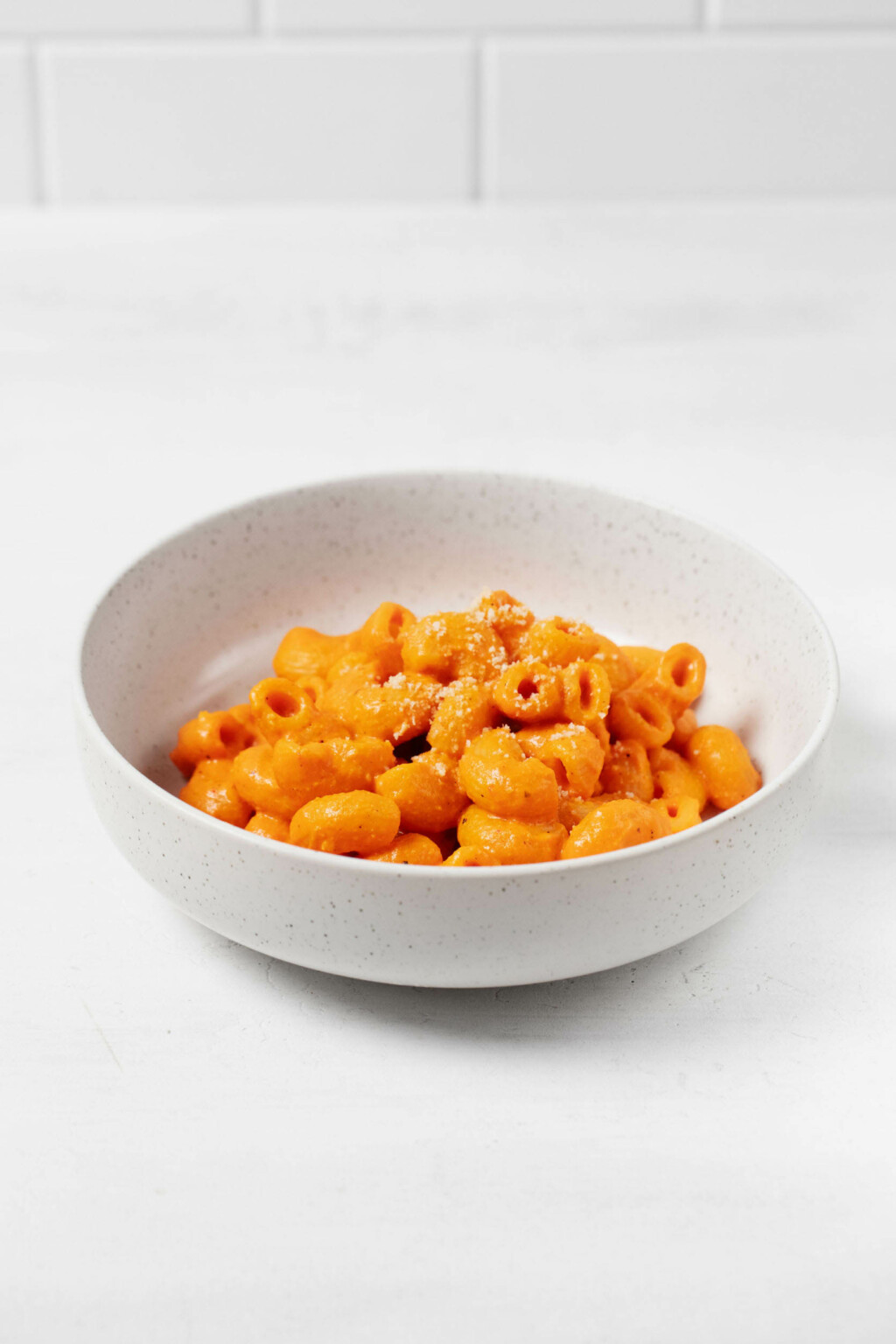 An angled photograph of a vegan red pepper mac and cheese, which is served in a round, white ceramic bowl. The bowl is resting on a white surface.