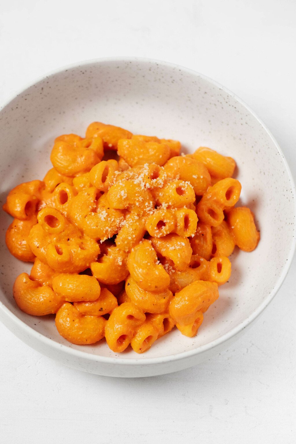 A round, white ceramic bowl has been filled with a plant-based mac and cheese. The mac and cheese has red peppers in the sauce, so it has a lovely orange color.