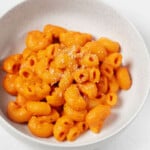 A round, white ceramic bowl has been filled with a plant-based mac and cheese. The mac and cheese has red peppers in the sauce, so it has a lovely orange color.