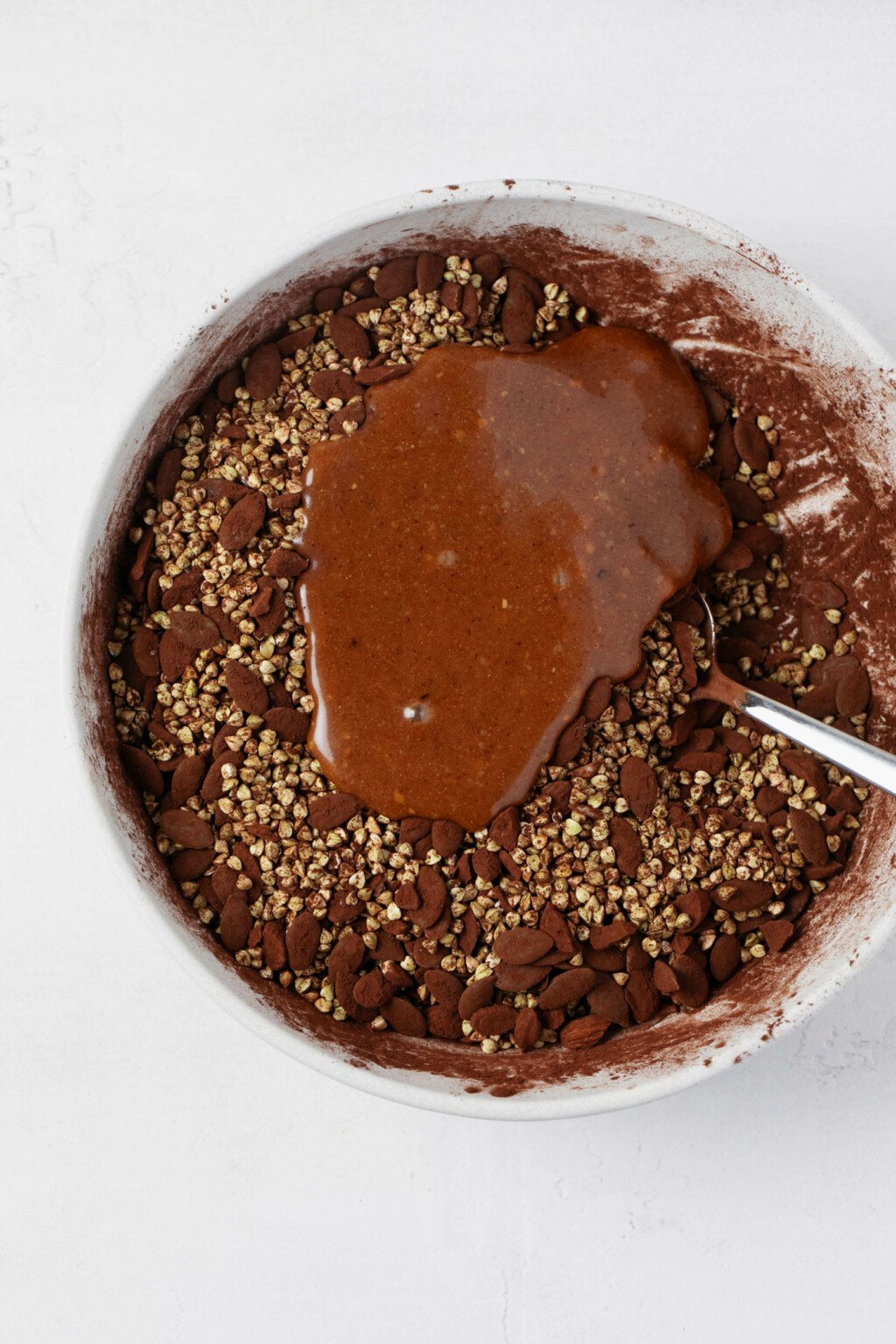 A mixing bowl is pictured overhead. It's filled with buckwheat groats and nut butter, along with cocoa powder.