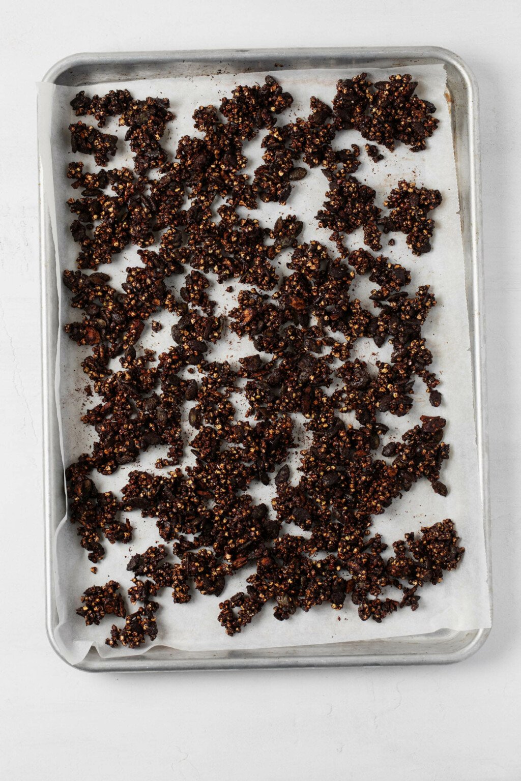 A baking sheet has been lined with parchment and is being used to bake cocoa-scented buckwheat granola clusters.