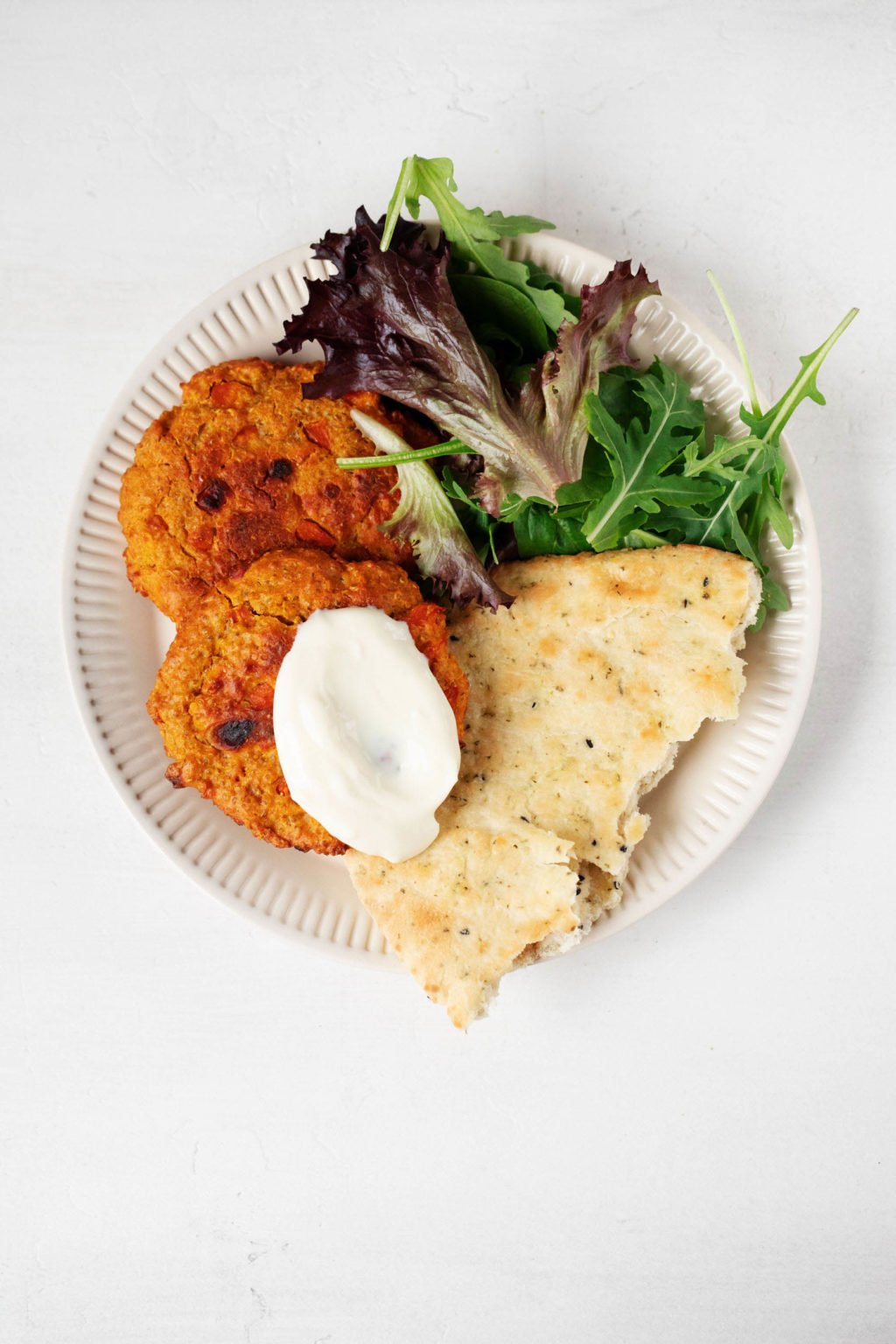 A white, rimmed plate has been topped with a piece of naan bread, mixed greens, plant-based patties, and a dollop of a dairy-free cream sauce. It rests on a white surface.