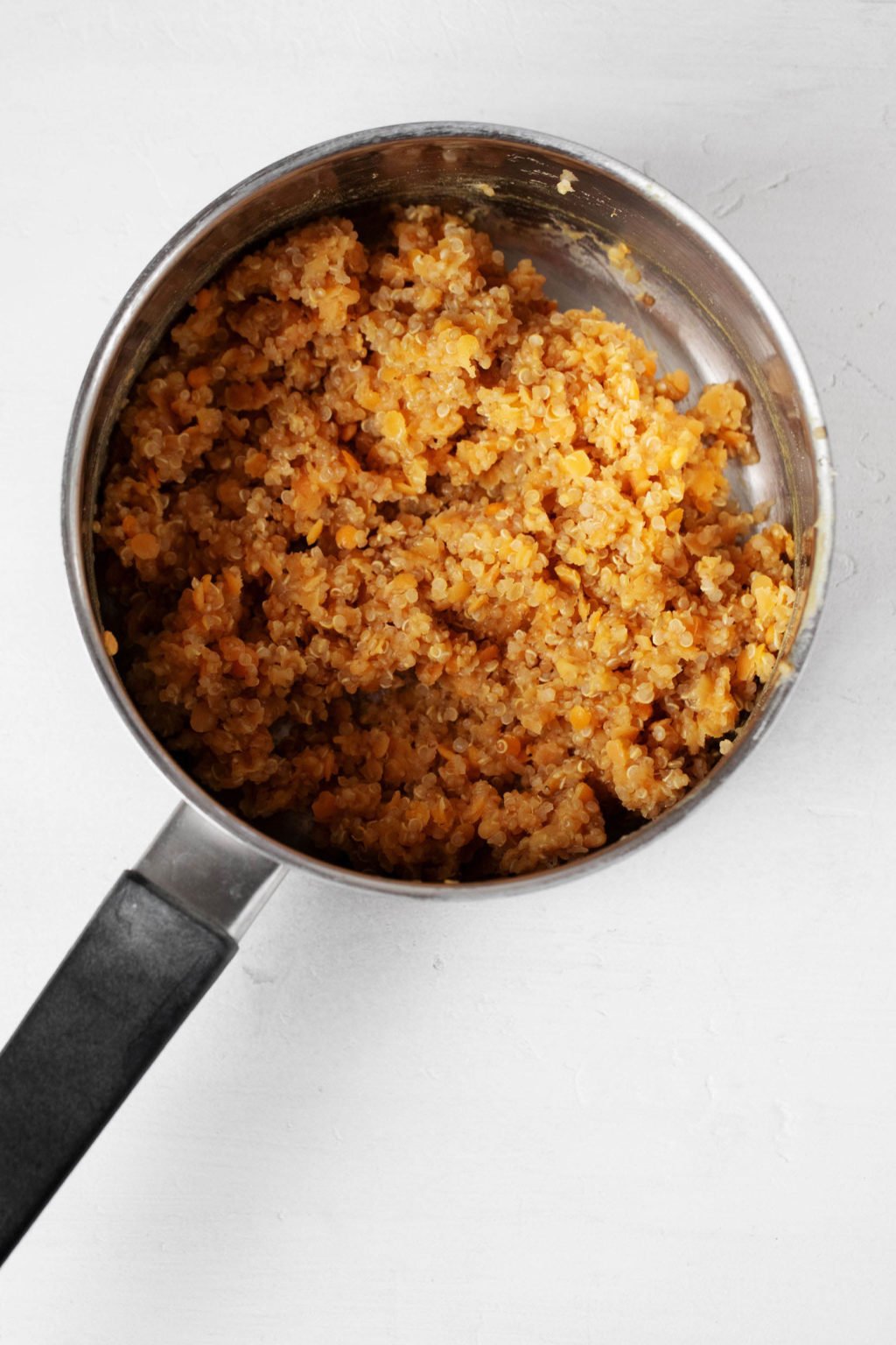 A silver saucepan is filled with a mixture of whole grains, legumes, onions, carrots, and celery. The mixture is a light orange color. The saucepan rests on a white surface.