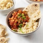 A tomato and chickpea based stew has been served over a bed of couscous, with a flatbread nearby and a garnish of green herbs.
