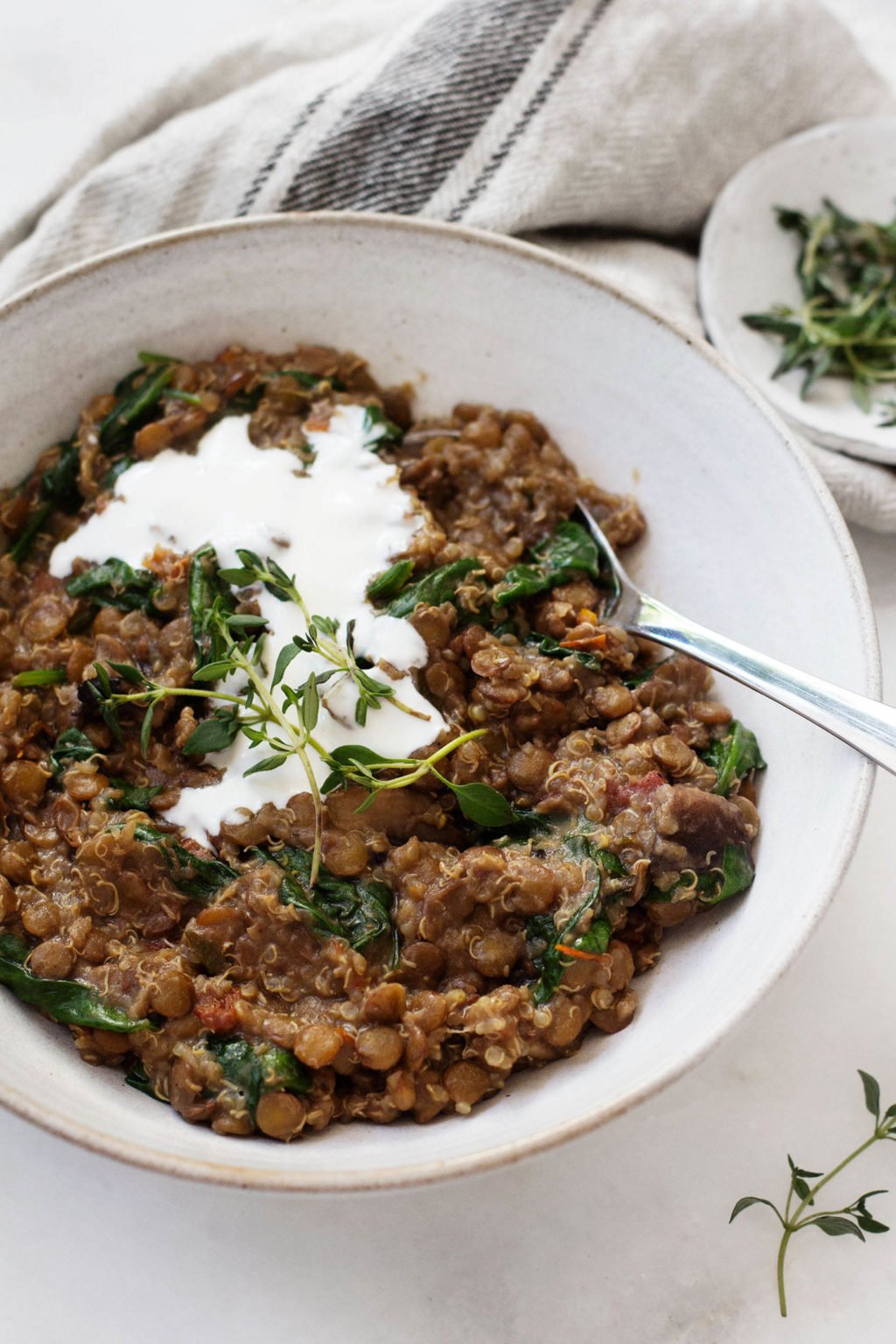 A bowl holding quinoa and lentils, topped with a white cashew cream, is placed next to a gray and white napkin and a small pinch pot of herbs.