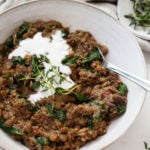 A bowl holding quinoa and lentils, topped with a white cashew cream, is placed next to a gray and white napkin and a small pinch pot of herbs.
