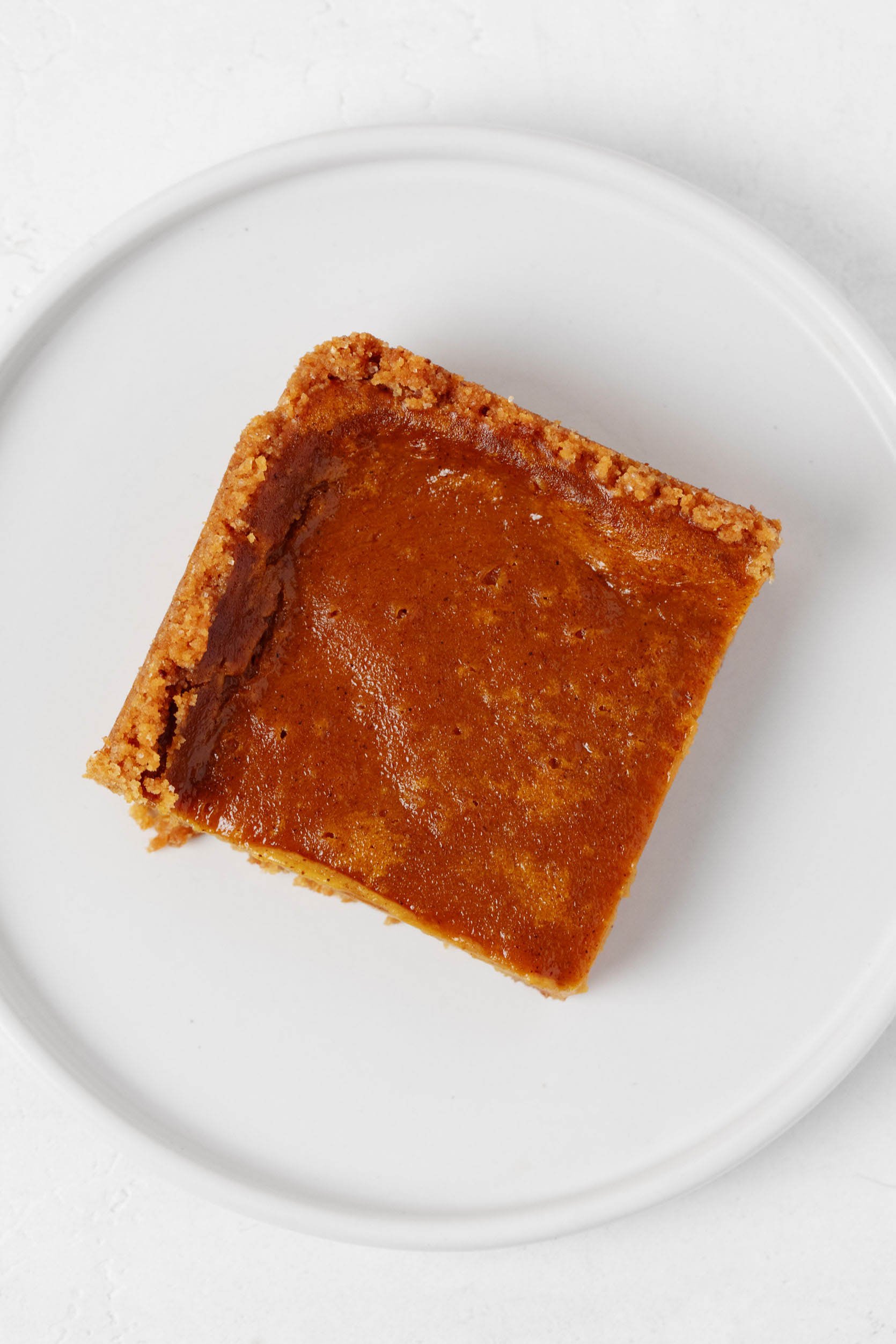 A perfectly square vegan pumpkin pie bar is pictured on a round, white plate.