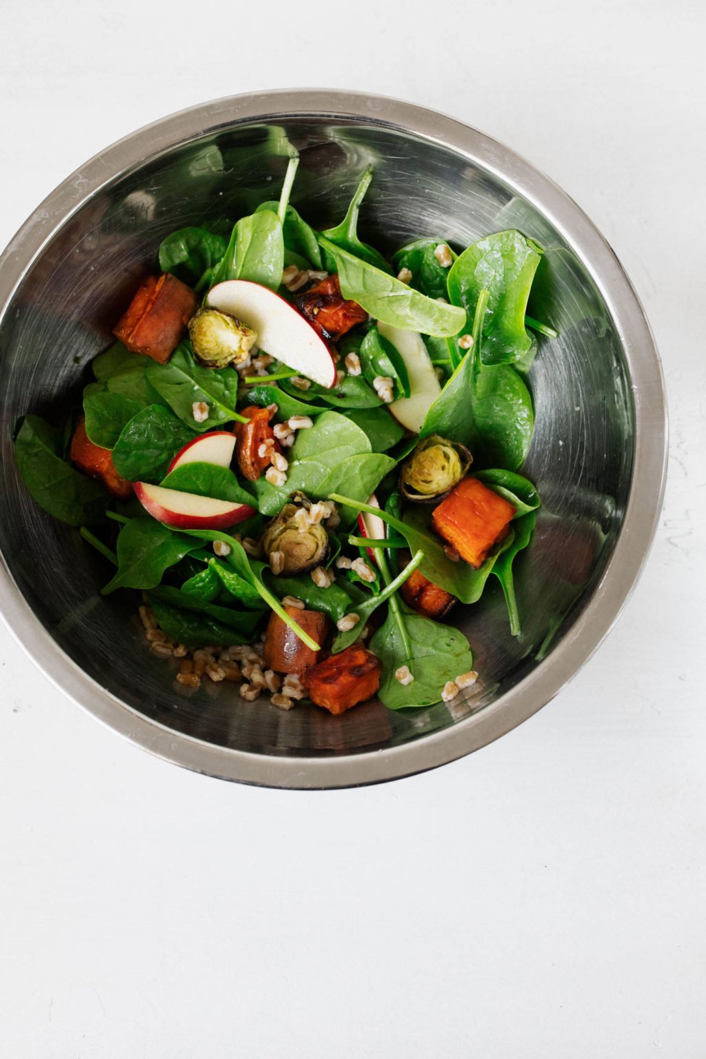 A stainless steel mixing bowl is full of the colorful ingredients for an autumn harvest salad. It rests on a white surface.