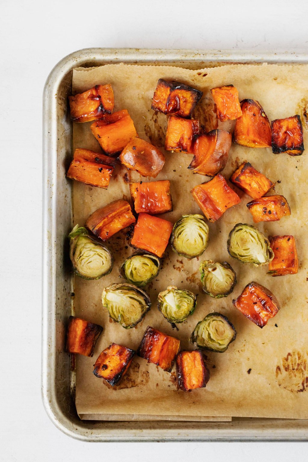 A baking sheet is covered in a layer of parchment, along with roasted sweet potatoes and Brussels sprouts.