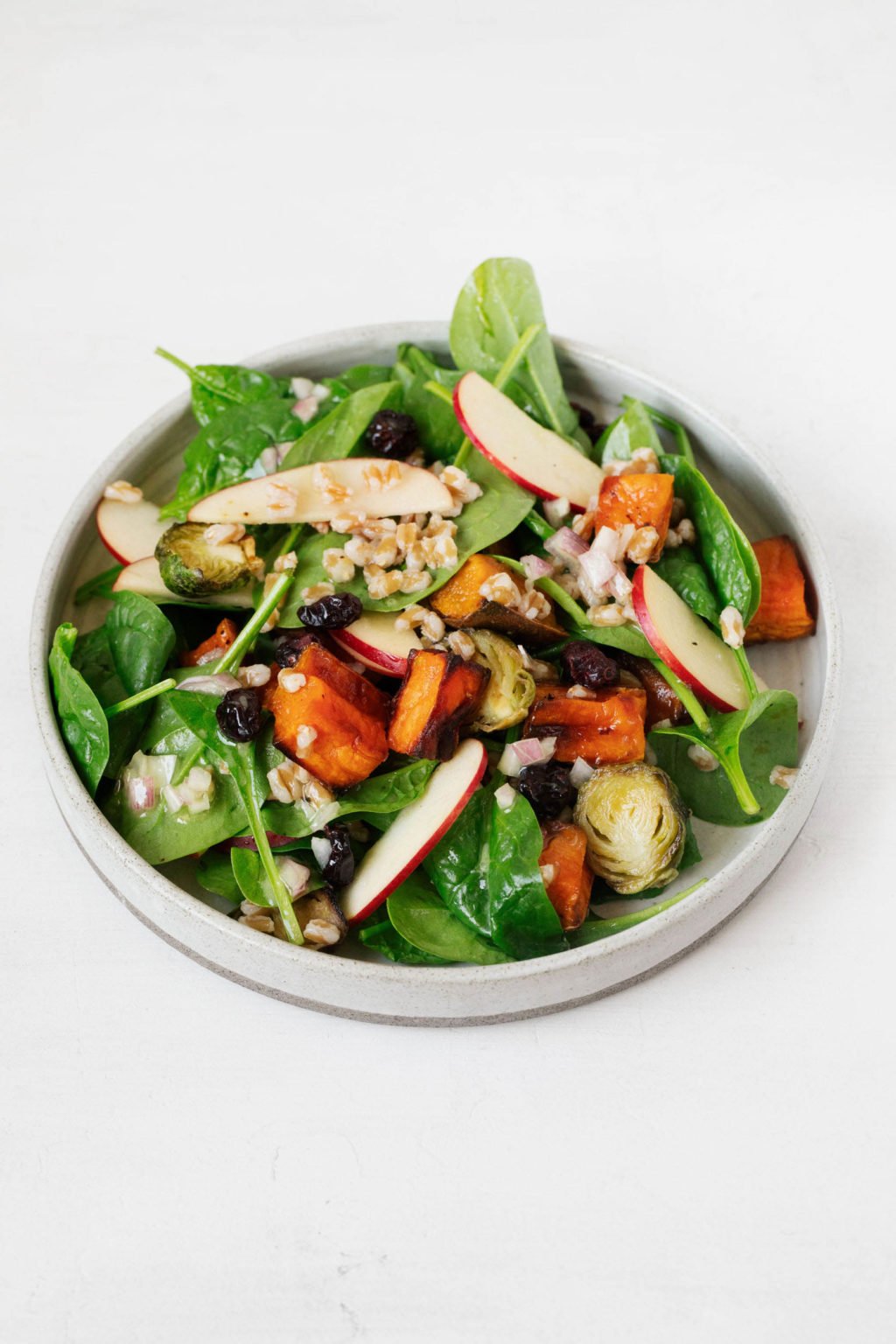 A rimmed, white salad plate is piled high with greens, roasted potatoes, thinly sliced apples, and cooked whole grains. Everything is dressed with a vinaigrette.