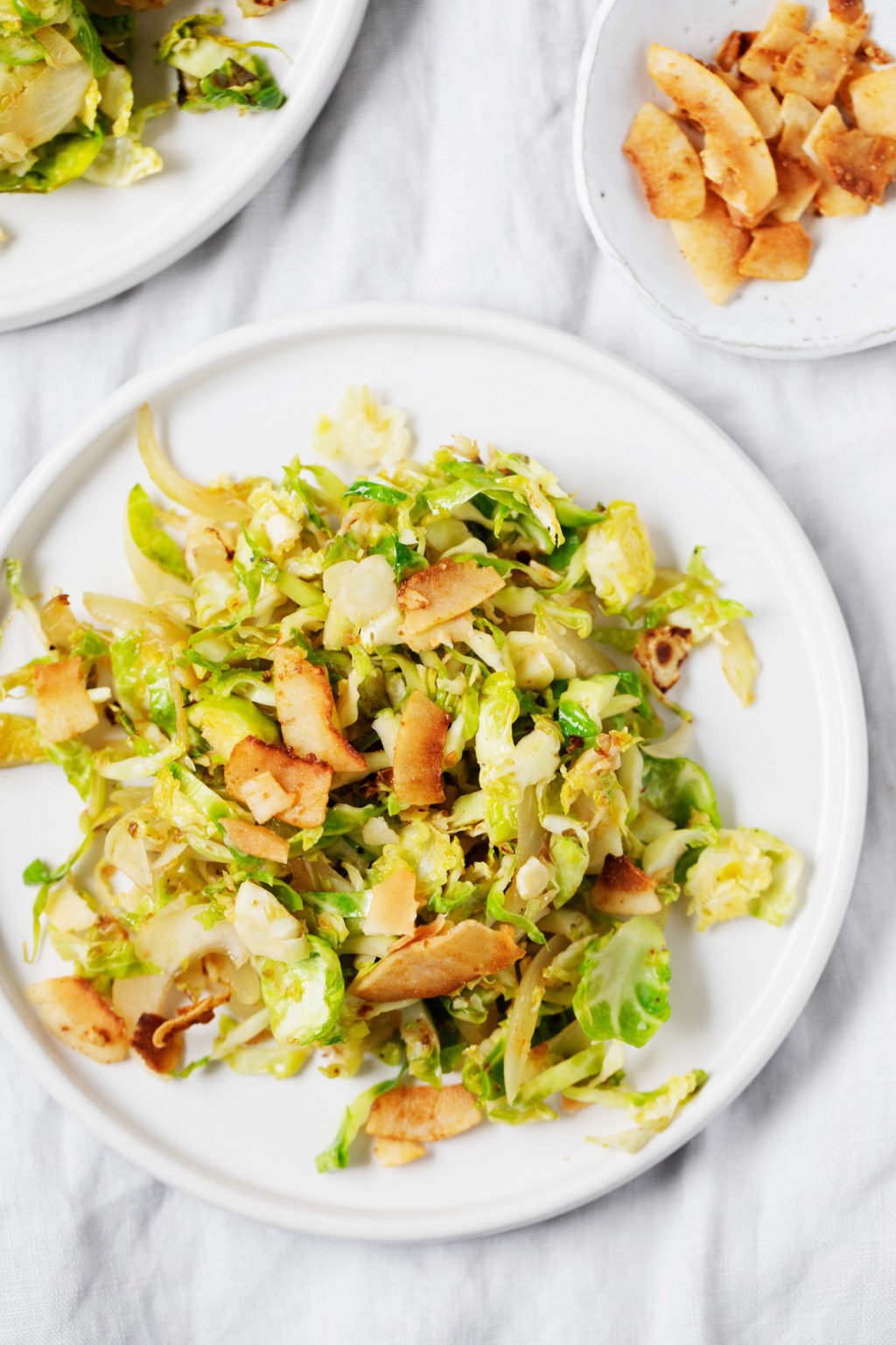 A white serving plate is topped with shaved Brussels sprout and a vegan bacon, which is made from seasoned coconut flakes.