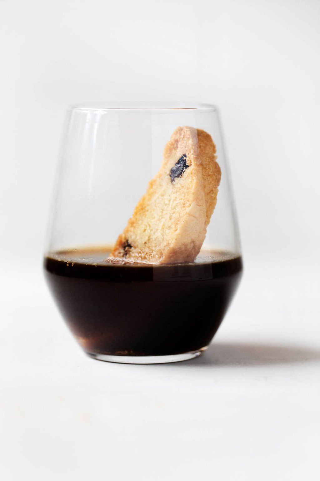 A clear glass of black coffee, with a piece of biscotti dunked inside.