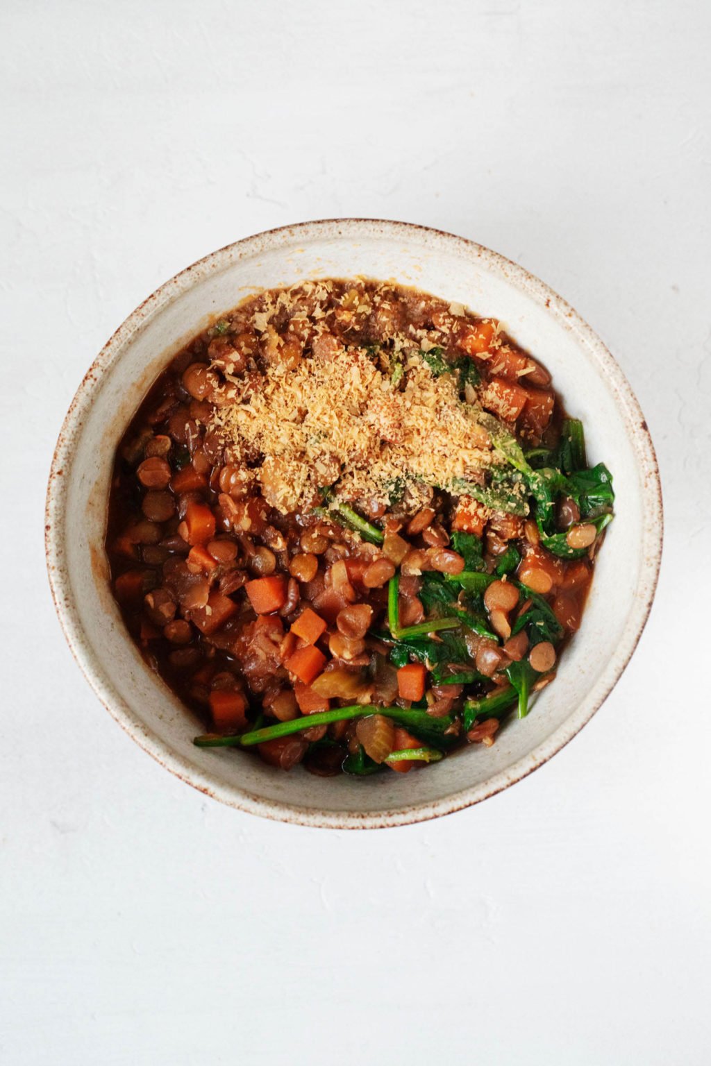 A round, cream colored bowl with a light brown rim holds a heaping cup of red brown lentil soup. Some spinach is wilted into the soup.