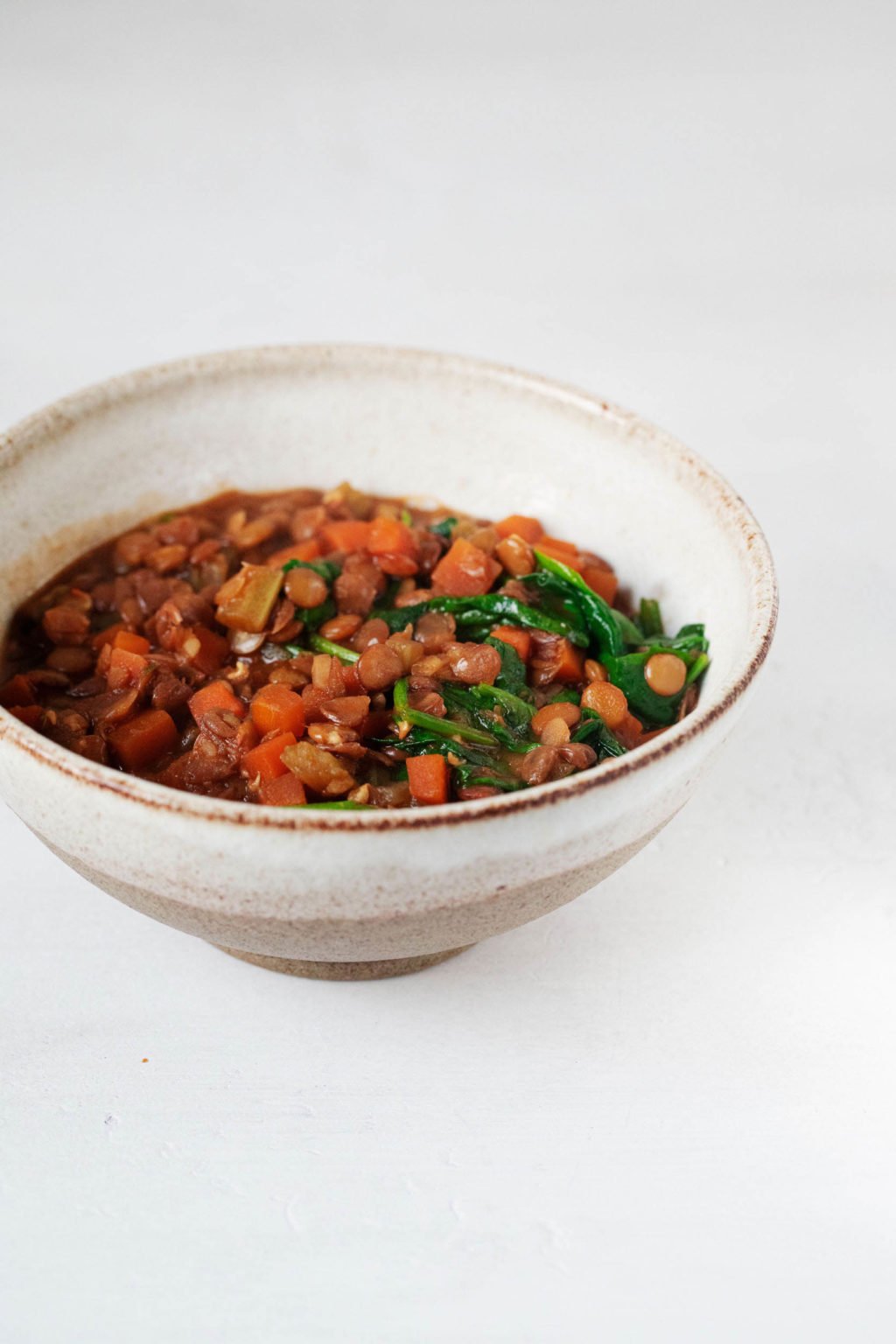 An angled photograph of a small, white bowl of vegetable and legume soup. It rests on a white surface.