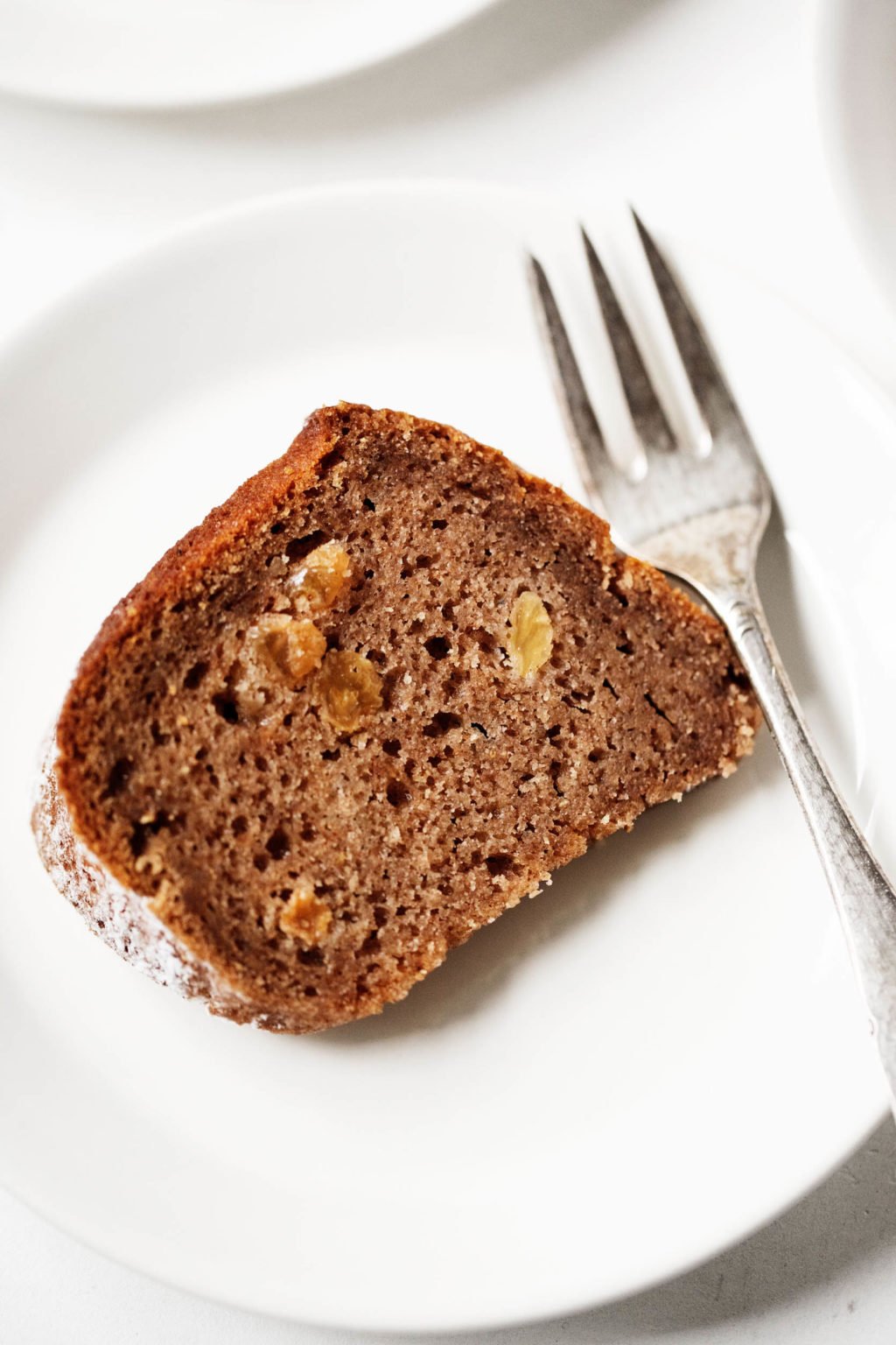 A white, ceramic dessert plate has been topped with a slice of vegan applesauce cake and is accompanied by a small, antique fork.