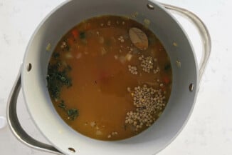 A gray soup pot is filled with broth, a bay leaf, and lentils.