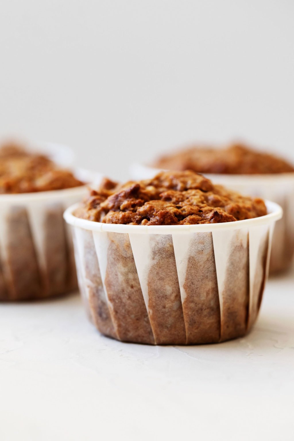 An angled photograph of vegan apple bran muffins, each wrapped in a white liner, resting on a white surface.