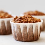 An angled photograph of vegan apple bran muffins, each wrapped in a white liner, resting on a white surface.