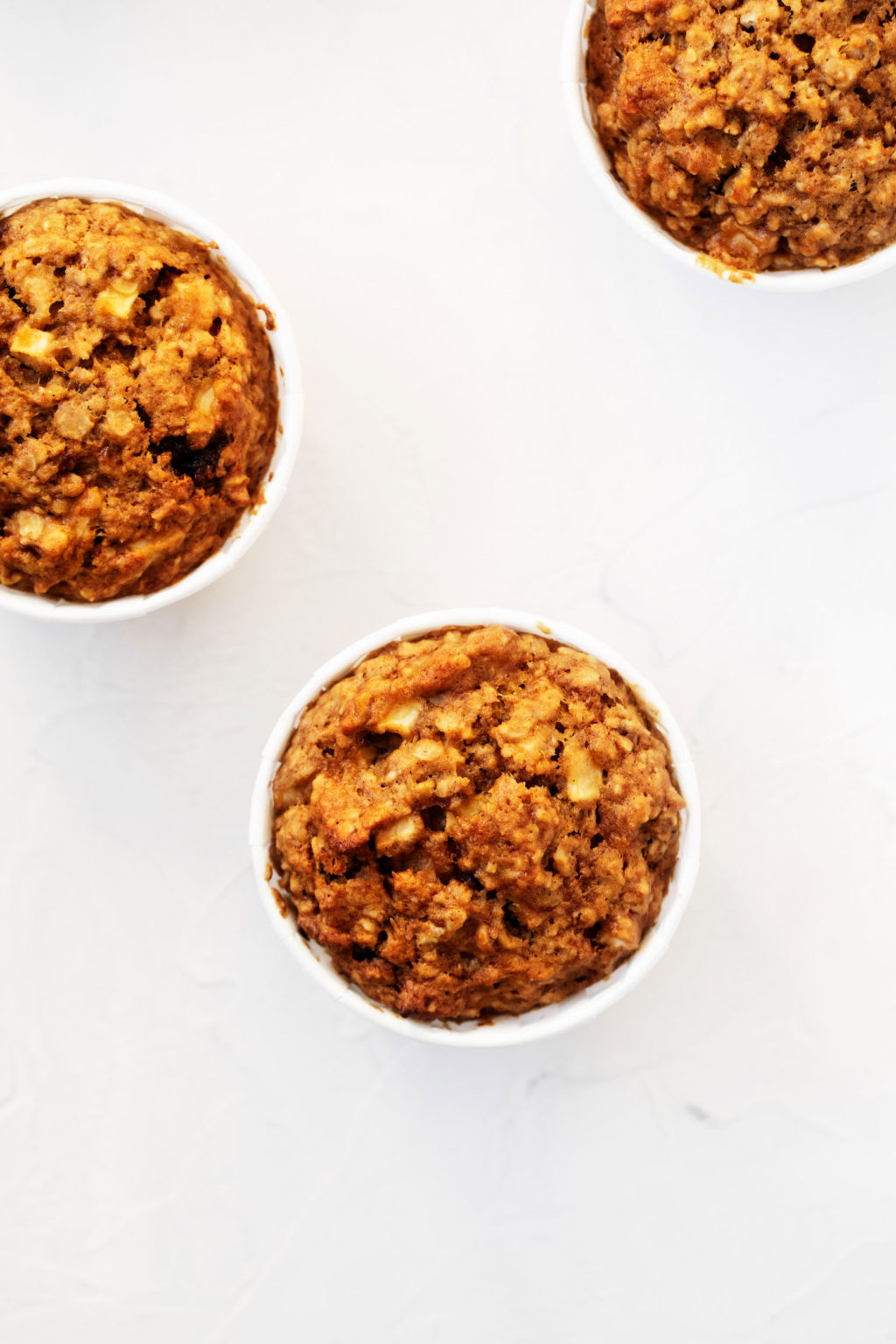 A few whole grain vegan muffins are resting on a white surface. Each is wrapped in a white liner.