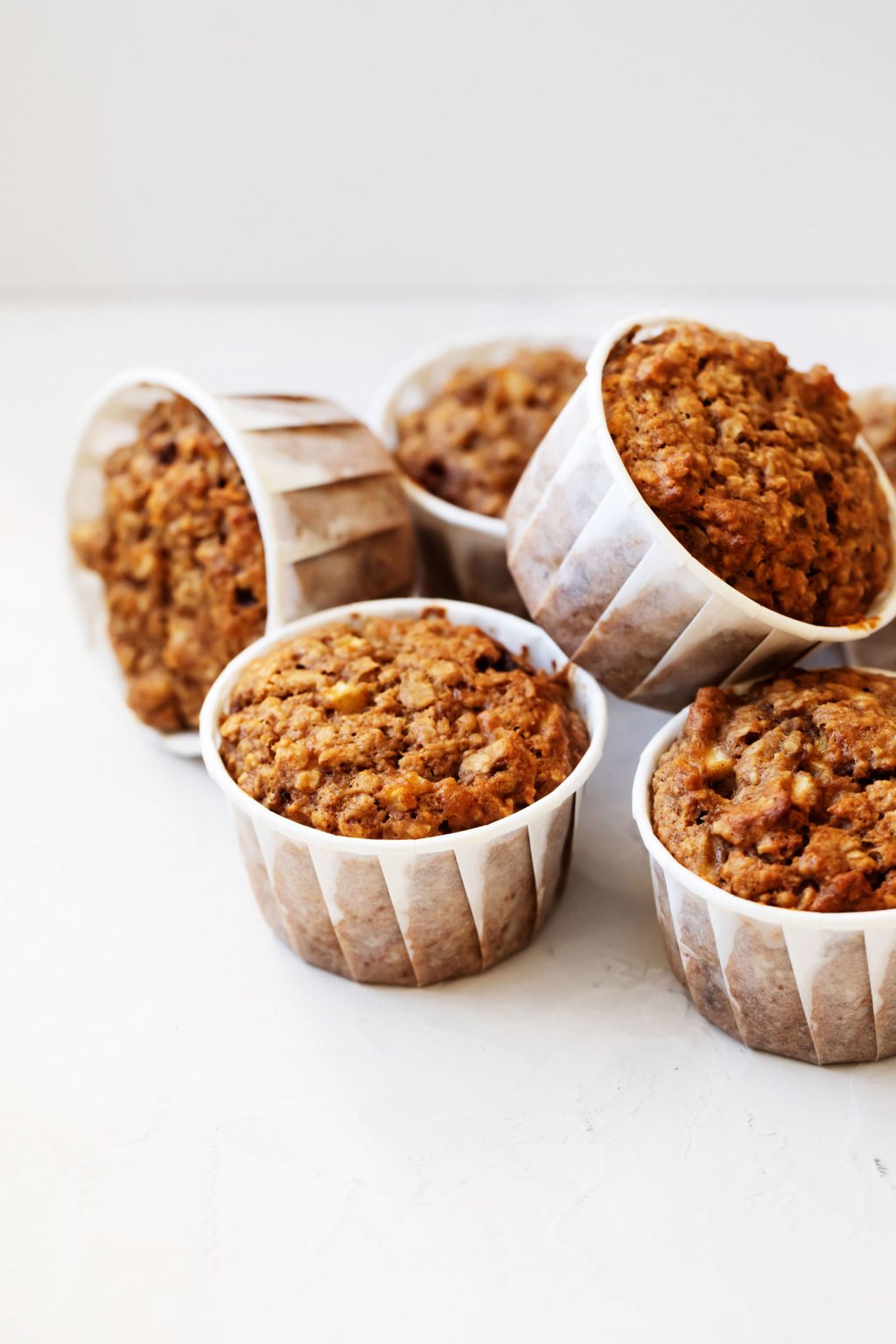 Six vegan apple bran muffins are stacked on a white surface. Some muffins are upright, while others lie sideways.