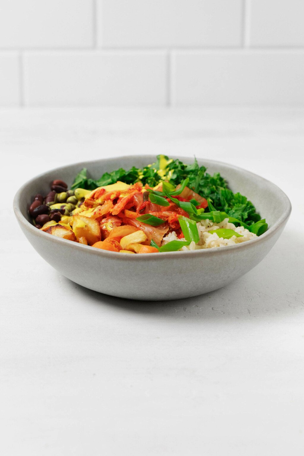 A gray bowl holds a mixture of whole grains, vegetables, beans, and a bright, yellow colored sauce. It's resting on a white surface with white tiles in the background.