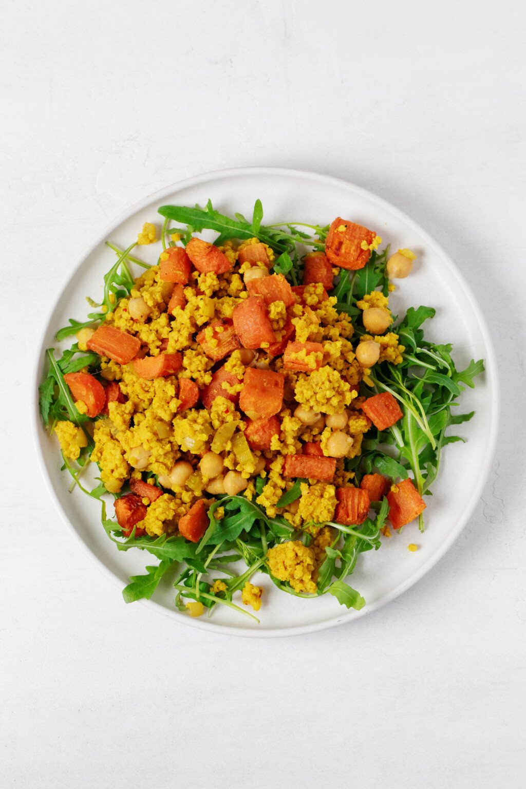 Greens, cooked whole grains, and roasted carrots are arranged on a round, white plate. 