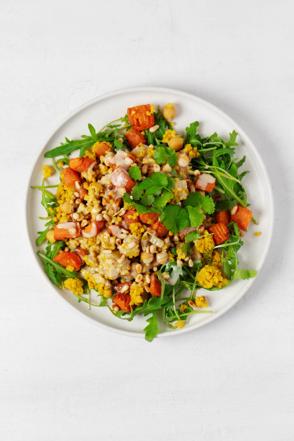 A round, white dish holds a golden-colored spiced millet pilaf. It's served with arugula, roasted carrots, and tahini dressing.