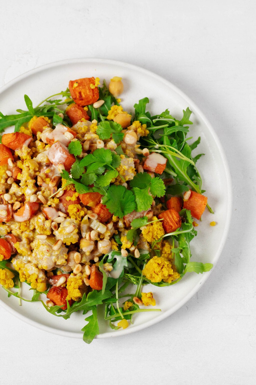 A round, white dish holds a golden-colored spiced millet pilaf. It's served with arugula, roasted carrots, and tahini dressing. 