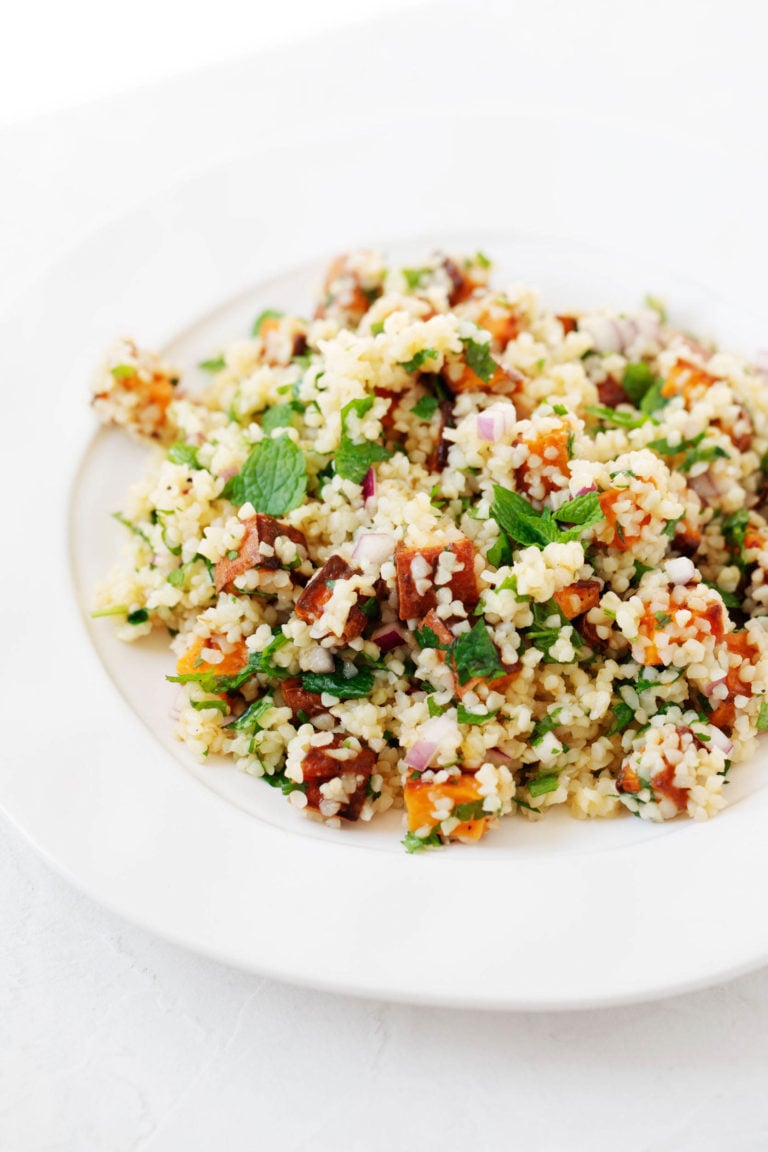 A wide-rimmed, white bowl is filled with a cooked vegan whole grain and sweet potato salad with herbs.
