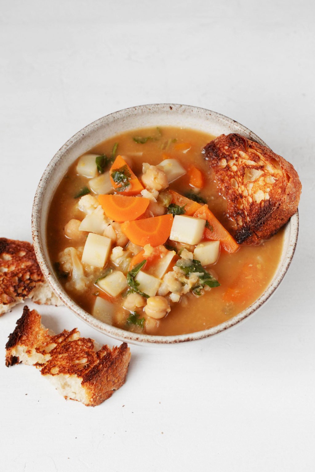 A soup bowl has been filled with a miso vegetable stew and is served with toasted bread. It sits on a white surface.