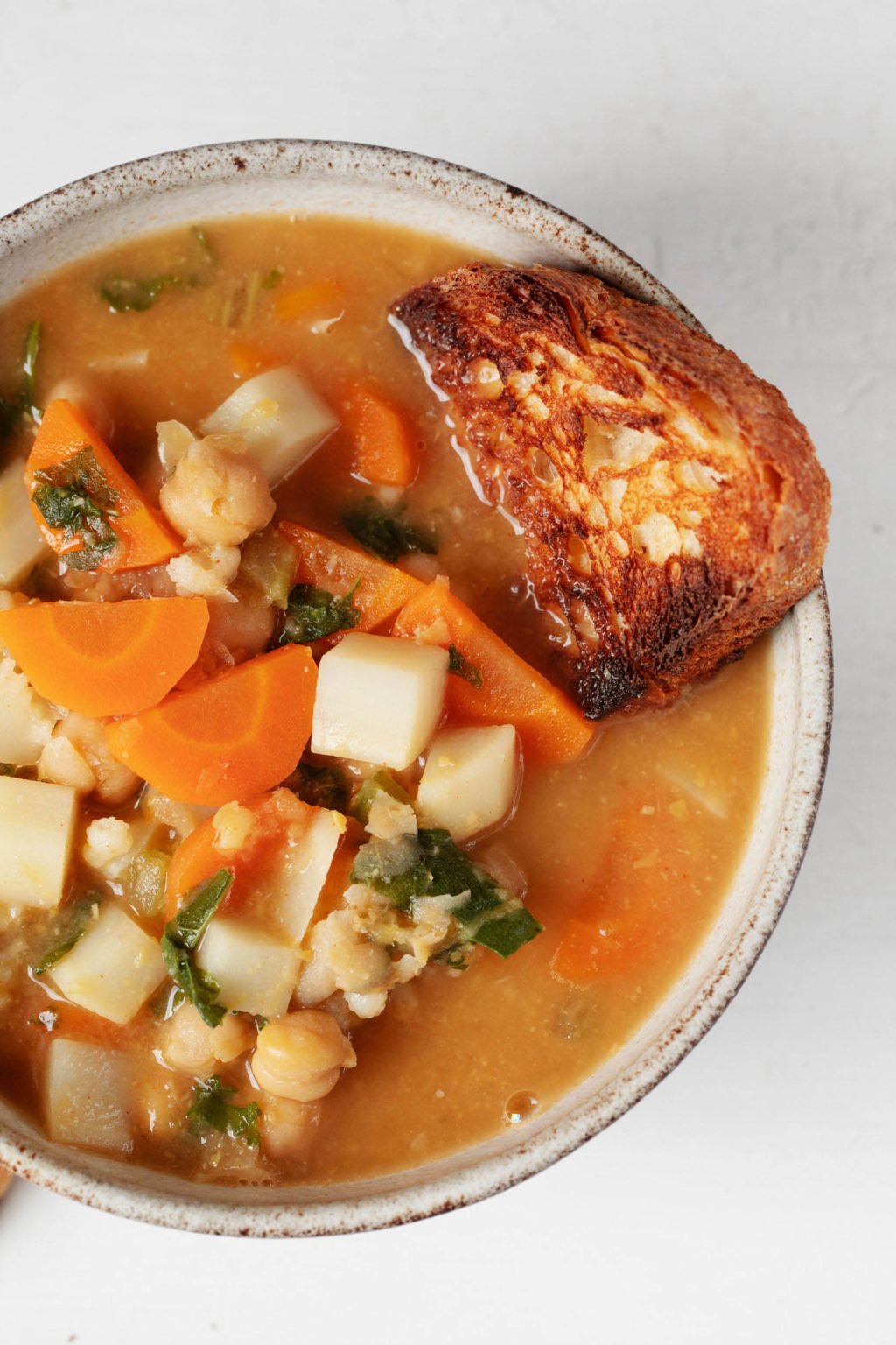 An overhead image of a hearty soup with carrots, chickpeas, and greens, which is served with a slice of toast.