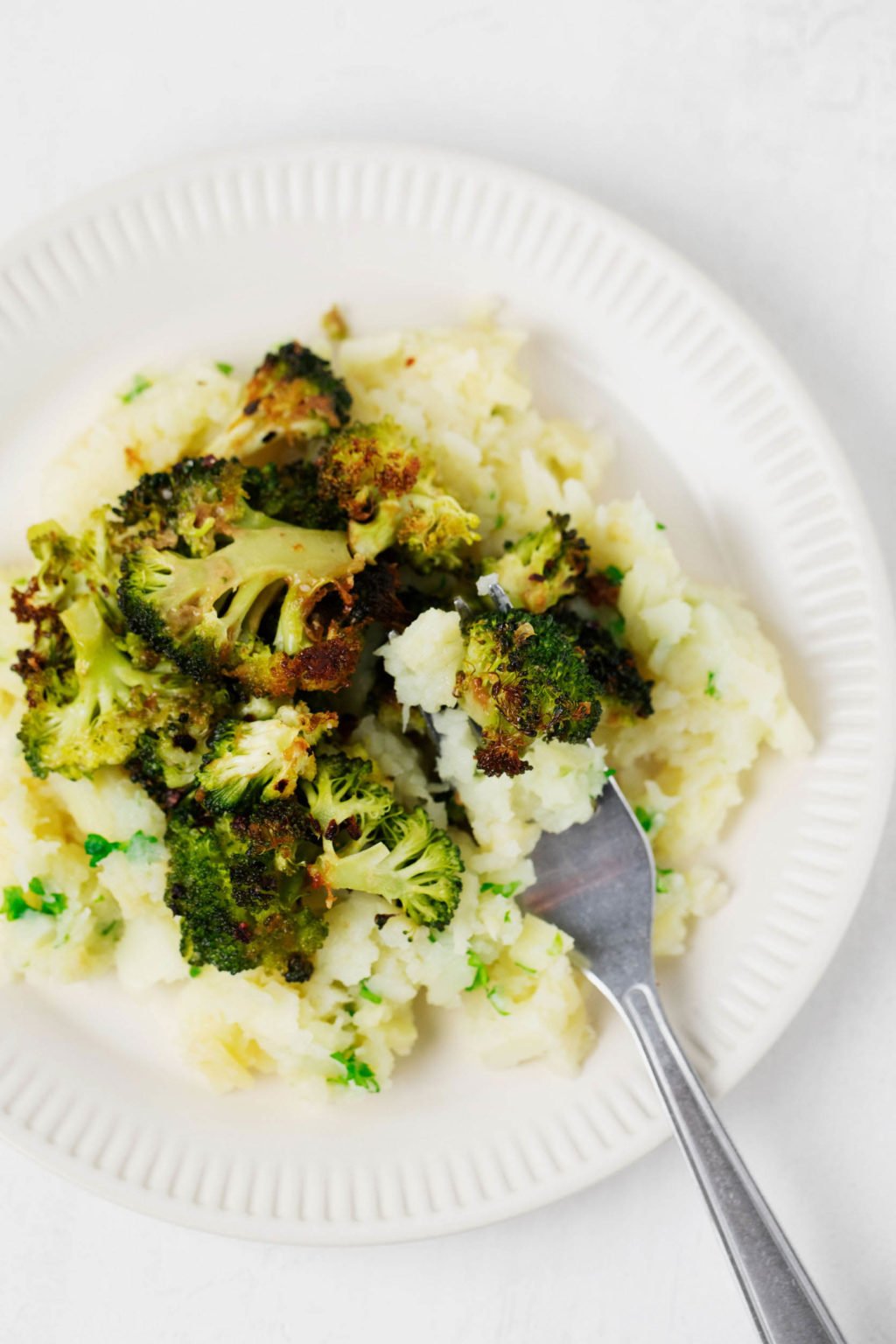 A mound of smashed root vegetables has been topped with crispy roasted broccoli and served on a white rimmed plate.