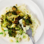 A mound of smashed root vegetables has been topped with crispy roasted broccoli and served on a white rimmed plate.