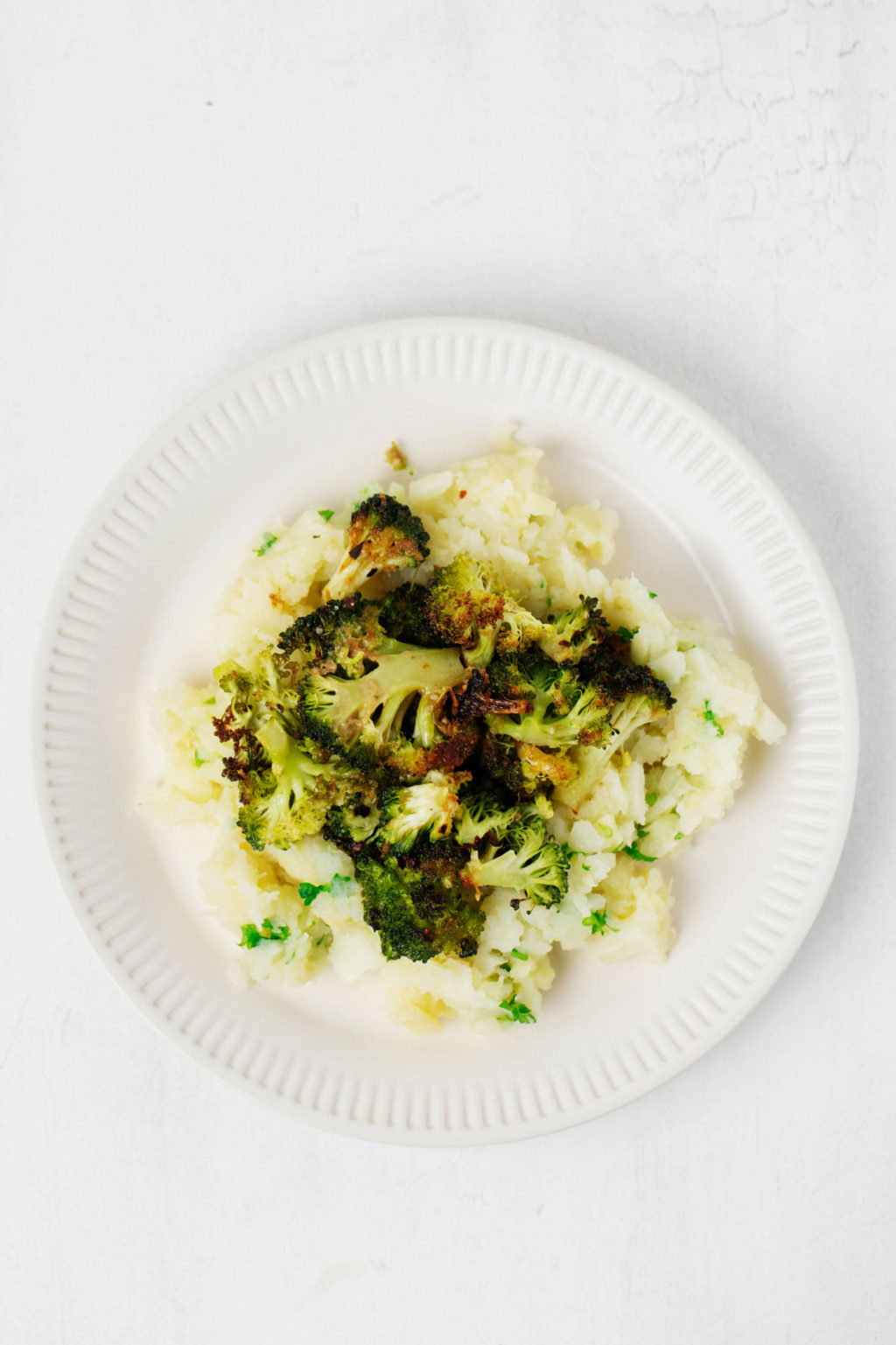 A white rimmed plate holds crispy roasted broccoli served over mashed root vegetables. It rests on a white surface.